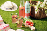 Outdoor picnic scene with Penrhos gin, glasses of pink cocktail, and a basket on grass.